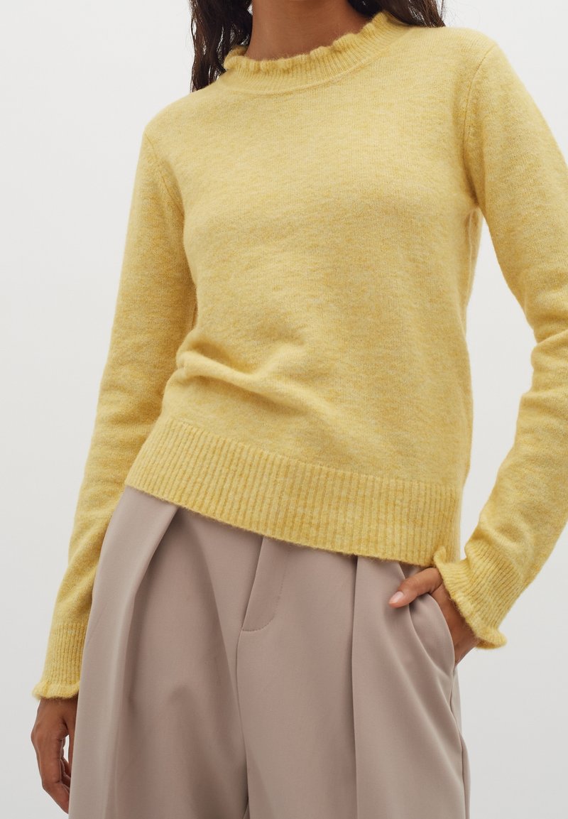 Femme portant un pull doux à manches longues jaune pâle associé à un pantalon beige taille haute, une main dans la poche et l'autre détendue.