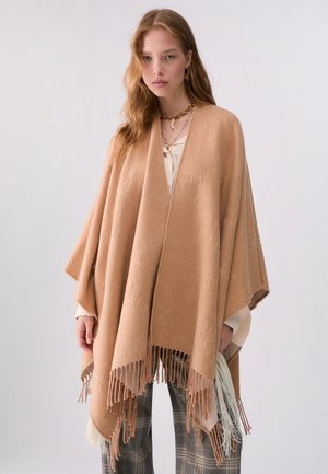 Jeune femme portant un poncho beige à franges par-dessus un chemisier blanc et un pantalon à carreaux, debout devant un fond clair uni.