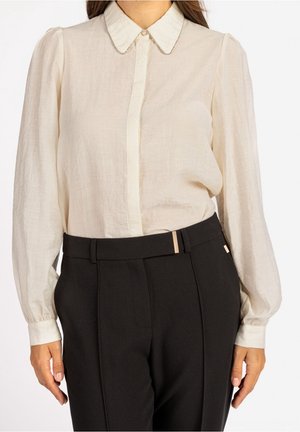 Femme portant une blouse beige clair à manches longues avec un col pointu et un pantalon noir taille haute avec un détail ceinture.