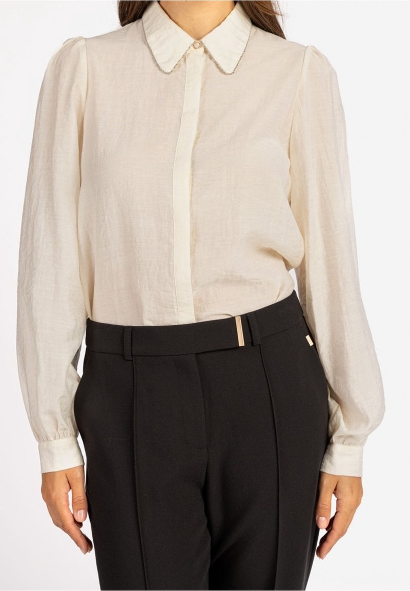Femme portant une blouse beige clair à manches longues avec un col pointu et un pantalon noir taille haute avec un détail ceinture.
