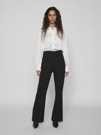 White ruffled blouse with long sleeves paired with high-waisted black flared pants. Smooth fabric and fitted design, featuring pointed black heels.