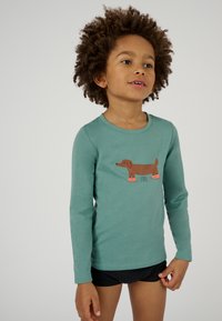 Maglietta a maniche lunghe color teal con una grafica di un bassotto marrone che indossa stivaletti arancioni. Tessuto morbido e scollatura rotonda. Indossata con pantaloni corti neri.