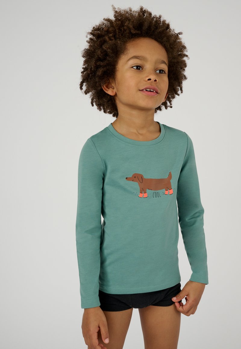 Maglietta a maniche lunghe color teal con una grafica di un bassotto marrone che indossa stivaletti arancioni. Tessuto morbido e scollatura rotonda. Indossata con pantaloni corti neri.