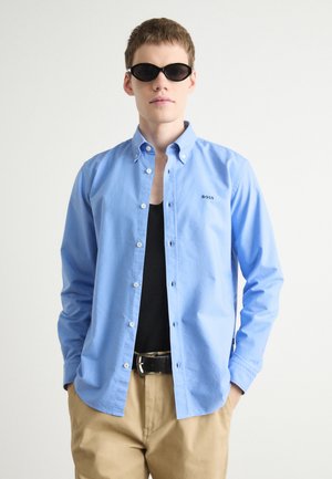Light blue button-up shirt with long sleeves and a small embroidered "BOSS" logo, worn over a black tank top and beige trousers.