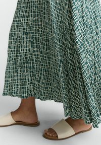 Jupe maxi verte à motifs avec un design géométrique blanc, associée à des sandales à glissière beiges sur un fond clair.