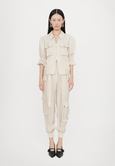 Beige kurze Jacke mit zwei vorderen Taschen und einem Reißverschluss, kombiniert mit passenden Cargo-Hosen mit Seitentaschen, abgerundet mit spitzen schwarzen Schuhen.