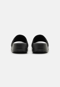 Chaussures noires avec une tige texturée, un dos ouvert et une semelle solide. Le design présente un style minimaliste et une finition lisse.