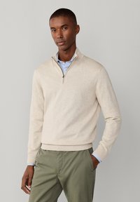 Mężczyzna w beżowym swetrze z zamkiem do połowy długości, nałożonym na jasnoniebieską koszulę i oliwkowo-zielone spodnie, stoi z jedną ręką w kieszeni na jednolitym szarym tle.