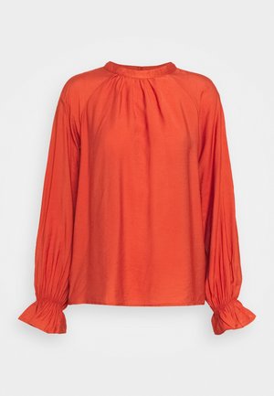 Blouse orange à manches longues, ample, avec un col froncé et des poignets plissés, présentée sur un fond clair et uni.
