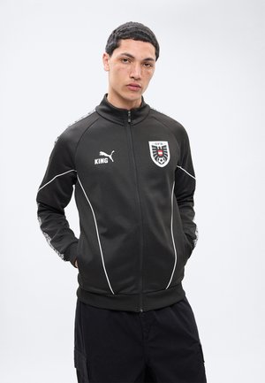 Junger Mann mit kurzen Haaren und Sommersprossen, der eine schwarze Puma-Trainingsjacke mit OFB-Fußballwappen und dem Schriftzug "KING" trägt, die Hände in den Taschen.