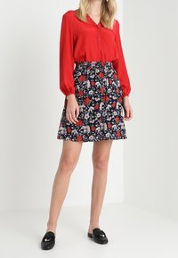 Punainen pitkähihainen pusero, jossa on nappidetaljit, yhdistettynä polvipituiseen tummakukkaiseen hameeseen, jossa on punaisia, valkoisia ja sinisiä kuvioita. Mustat loaferit.