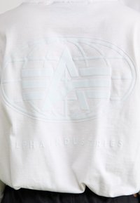 Hvit langermet skjorte med et stort, preget logo i lys blå farge på baksiden, med teksten "ALPHA INDUSTRIES" under grafikken.