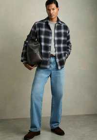 Karierte Flanellhemd in Marineblau und Grau, graues T-Shirt, hellblaue Jeans, braune Slipper und eine dunkle Ledertasche.