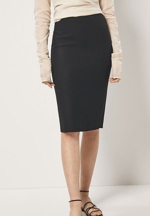 Kvinde iført sort knælang pencilnederdel, beige langærmet gennemsigtig top og sorte sandaler med stropper, stående mod en ensfarvet baggrund.