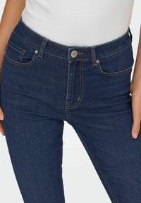 Granatowe dżinsy o wysokim stanie, wyposażone w przednie kieszenie, metalowe zapięcie na guzik oraz widoczne akcenty ściegów. Gładka struktura.
