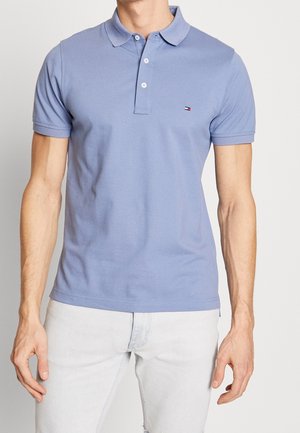 Homme portant un polo à manches courtes bleu clair avec des boutons et un petit logo sur la poitrine, associé à un pantalon de couleur claire, debout devant un fond blanc.