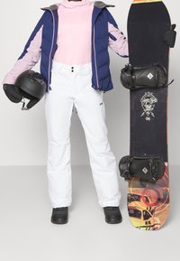 Person wearing a blue and pink jacket, pink turtleneck, white snow pants, black gloves holding helmet, and black boots next to a snowboard.