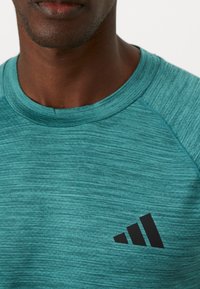 Türkiissinine sportlik särk, tekstuurse kangaga, �ümar kaelus ja must Adidas logo vasakul rinnal.