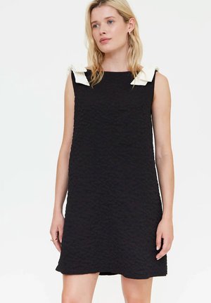 Mujer con vestido negro texturizado sin mangas con detalles de lazo blanco en los hombros, de pie frente a un fondo blanco liso.