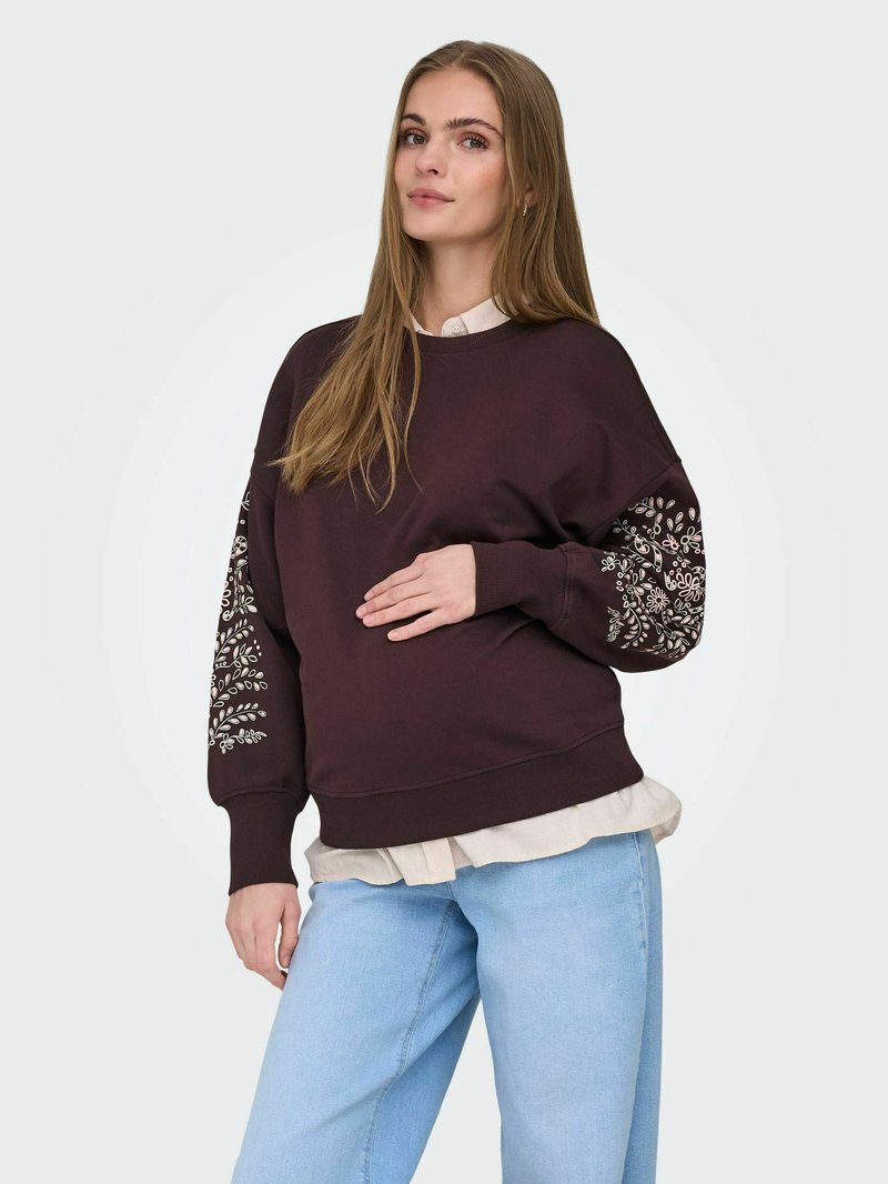 Woman with long hair wearing a dark brown sweatshirt with white floral embroidery on sleeves, light blue jeans, and a white shirt underneath.