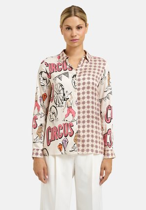 Frau trägt ein langärmeliges Hemd mit Knöpfen, das Zirkus- und geometrische Muster in Pink, Schwarz und Beige zeigt, kombiniert mit einer weißen Hose.
