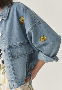 Denimjack, lichtblauw, ontspannen pasvorm. Voorzien van twee geborduurde bijenpatches op de mouwen, een zak aan de linkerkant en zilveren drukknopen.