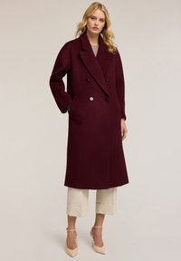 Cappotto in misto lana borgogna con design doppiopetto, bottoni neri e tasche laterali. Indossato sopra pantaloni cropped crema e tacchi nudi.