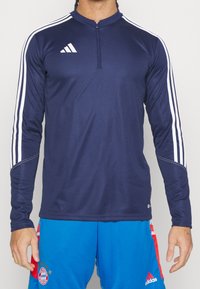 Muž v námořnickém dlouhém rukávu zipové mikiny Adidas s bílými pruhy a modrých šortkách FC Bayern Mnichov s červenými a bílými detaily.