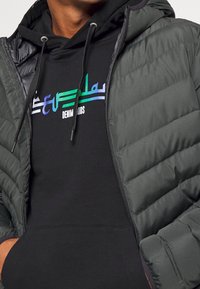 Chaqueta acolchada gris con secciones acolchadas y cierre de cremallera, sobrepuesta a una sudadera con capucha negra con un logo multicolor y cordones ajustables.