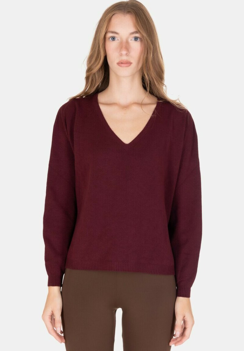 Donna con capelli castano chiaro lunghi che indossa un maglione a V color bordeaux e pantaloni marroni, rivolta verso l'obiettivo su uno sfondo bianco uniforme.