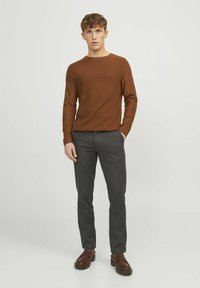 Brauner gestreifter Langarmpullover, graue maßgeschneiderte Hose und braune Slipper, vor einem hellen Hintergrund.