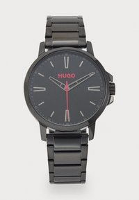 Montre en acier inoxydable noir avec un cadran rond. Le cadran noir présente des index horaires en argent, le nom de marque rouge "HUGO" et une aiguille des secondes rouge.