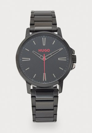Montre en acier inoxydable noir avec un cadran rond. Le cadran noir présente des index horaires en argent, le nom de marque rouge "HUGO" et une aiguille des secondes rouge.