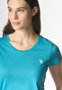 Una donna con capelli castani di lunghezza media indossa una T-shirt turchese brillante con un piccolo logo ricamato sul petto sinistro.