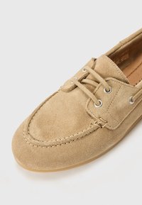 Beige selvskinnbåtssko med kontrasterende hvite sømmer, rund tå og metalløyelukker for snøring. Myk tekstur og lav profil.
