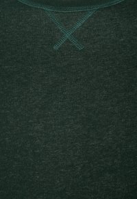 Tessuto verde scuro in misto cotone con una texture sottile. Presenta un design a punto croce sulla scollatura e un colletto a coste.