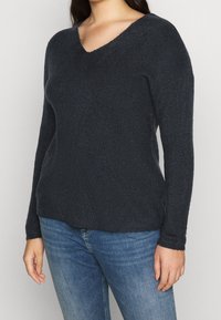 Dark navy V-neck sweater made of textured knit fabric, long sleeves, and a relaxed fit. Worn with blue jeans, showing casual layering.
