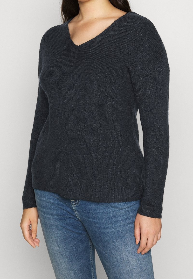 Dark navy V-neck sweater made of textured knit fabric, long sleeves, and a relaxed fit. Worn with blue jeans, showing casual layering.