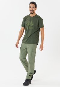 Olivgrön t-shirt med cykelgrafik, kombinerad med ljusgröna cargobyxor med fickor, som bärs med svarta skor.