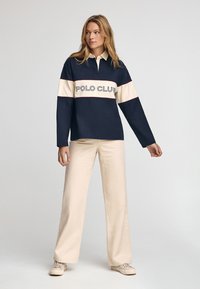Langarm-Polohemd in Marineblau mit cremefarbenem horizontalem Streifen und "POLO CLUB" Schriftzug. Kombiniert mit leichten, weit geschnittenen Hosen und beigen Schuhen.