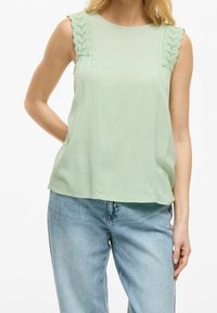 Miętowozielony top bez rękawów z teksturowaną koronkową lamówką na ramionach, miękki materiał, luźny krój i kieszenie po bokach, zestawiony z jasnoniebieskimi dżinsami.