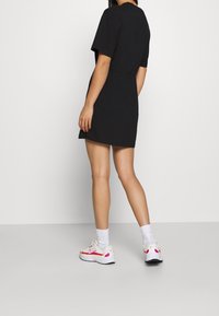 Svart kortärmad klänning med en åtstramad midja, kombinerad med vita sneakers med rosa detaljer och ankelsockor, visad bakifrån.