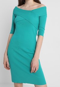 Türkises Off-Shoulder-Kleid mit Dreiviertelärmeln, tailliertem Design, strukturiertem Stoff, mit einem Kreuz-Frontdetail und knielangem Saum.