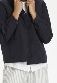Giacca corta navy con maniche lunghe, chiusura con bottoni e due tasche frontali, realizzata in tessuto liscio. Indossata sopra una camicia bianca e pantaloni a righe.