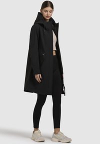 Manteau noir imperméable avec capuche, doté de poches latérales et d'une coupe décontractée, associé à des leggings noirs et des sneakers de couleur claire.