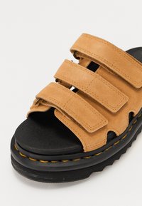 Sandal in suede marrone, con tre cinturini regolabili, suola in gomma nera e dettagli di cucitura gialla lungo il bordo e la suola.