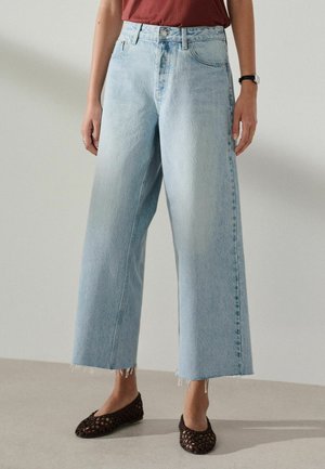 Persoană purtând blugi largi, albaștri deschiși, croiți scurt cu talie înaltă și tivuri destrămate, balerini maro împletiți, top roșu și un ceas de mână negru.