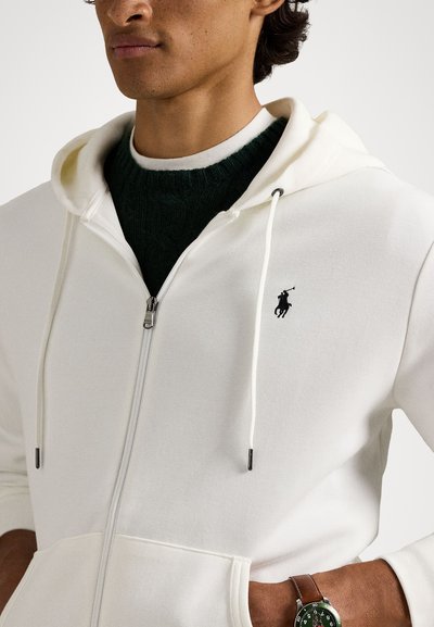 Fehér zipzáras kapucnis pulóver, zsinórokkal, egy kis fekete polo logóval. Sötétzöld pulóver felett viselve, látható egy barna óra.