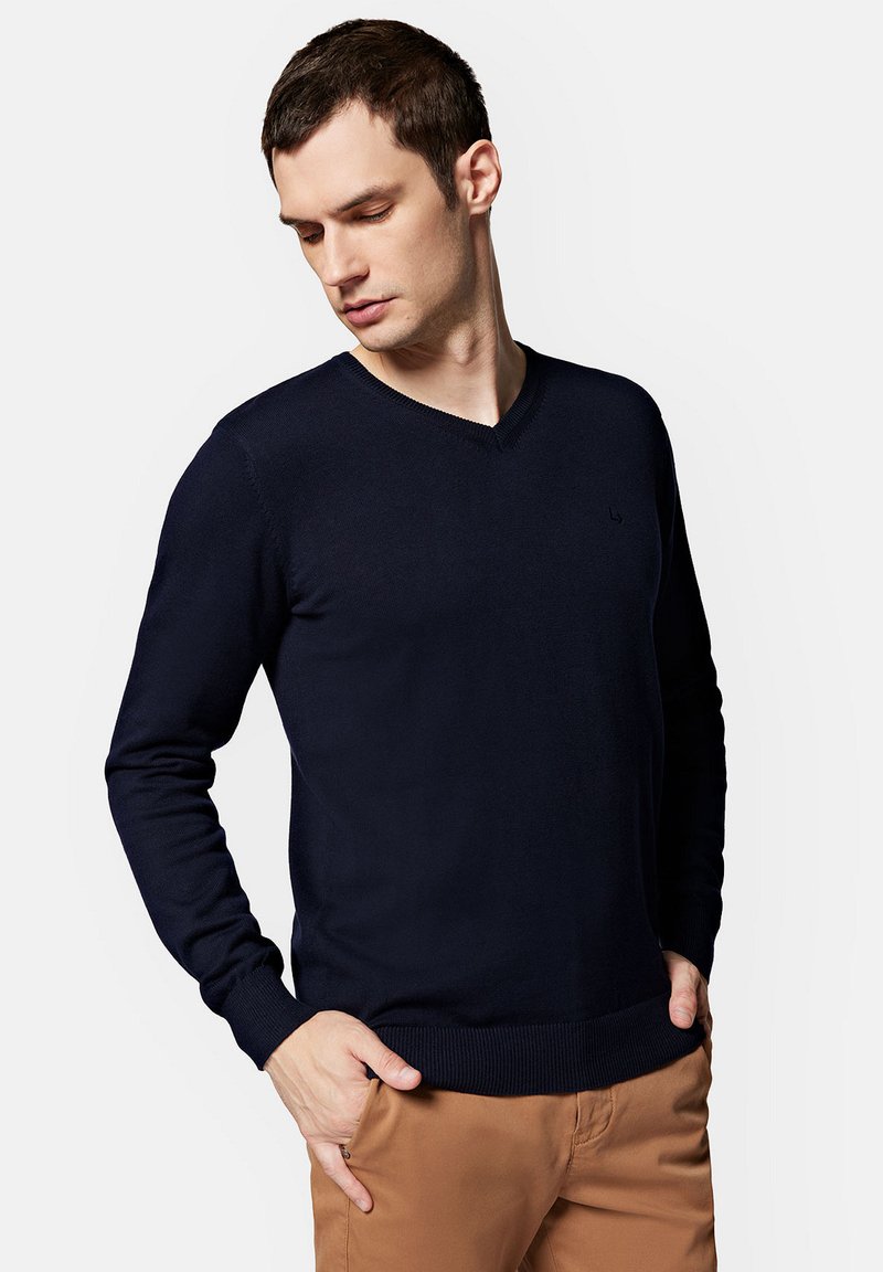 Granatowy sweter typu V-neck, wykonany z cienkiego przędzy, o dopasowanym kroju. Posiada ściągacze przy mankietach i u dołu, a na piersi znajduje się subtelny detal z logo.