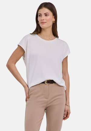 Frau mit braunen Haaren, die eine weiße Bluse mit kurzen Ärmeln, beigefarbene Hosen mit einem dunklen Gürtel, goldene Creolen und ein Armband trägt, posiert mit der Hand in der Hüfte.